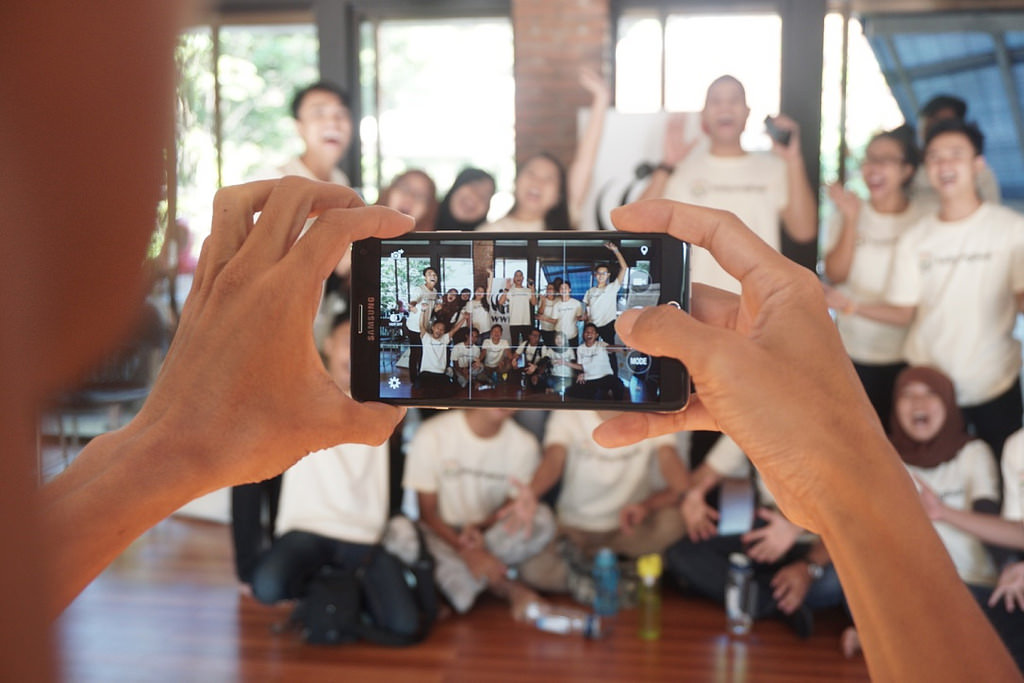 A group of people pose smiling for someone taking a photo on their phone.