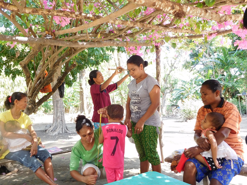 A woman measures the height of a toddler under a tree, surrounded by other women and children. (Vita Febriany)