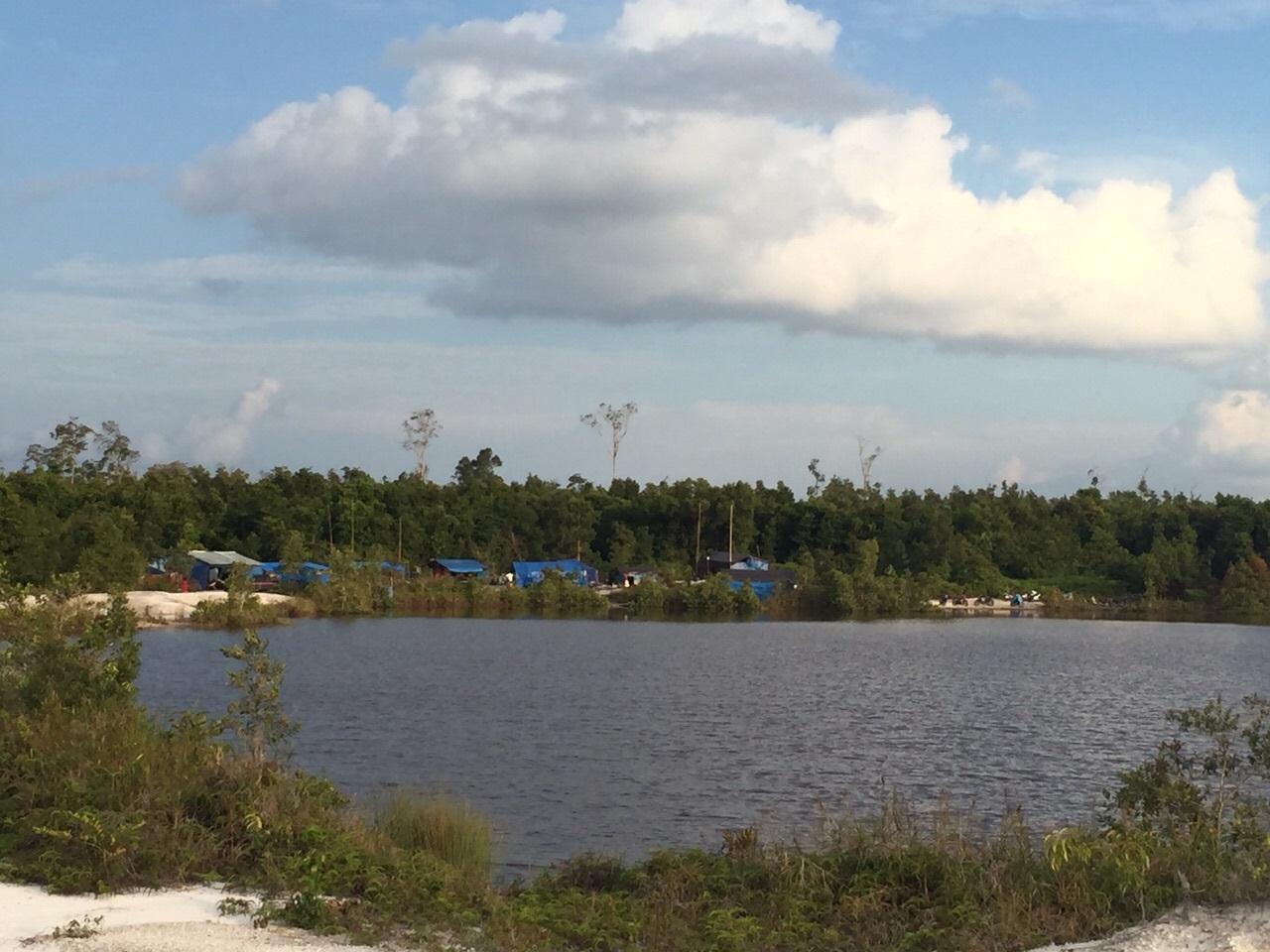 A view of a mine site with miners' tents in the background, next to a body of water but surrounded by barren land.