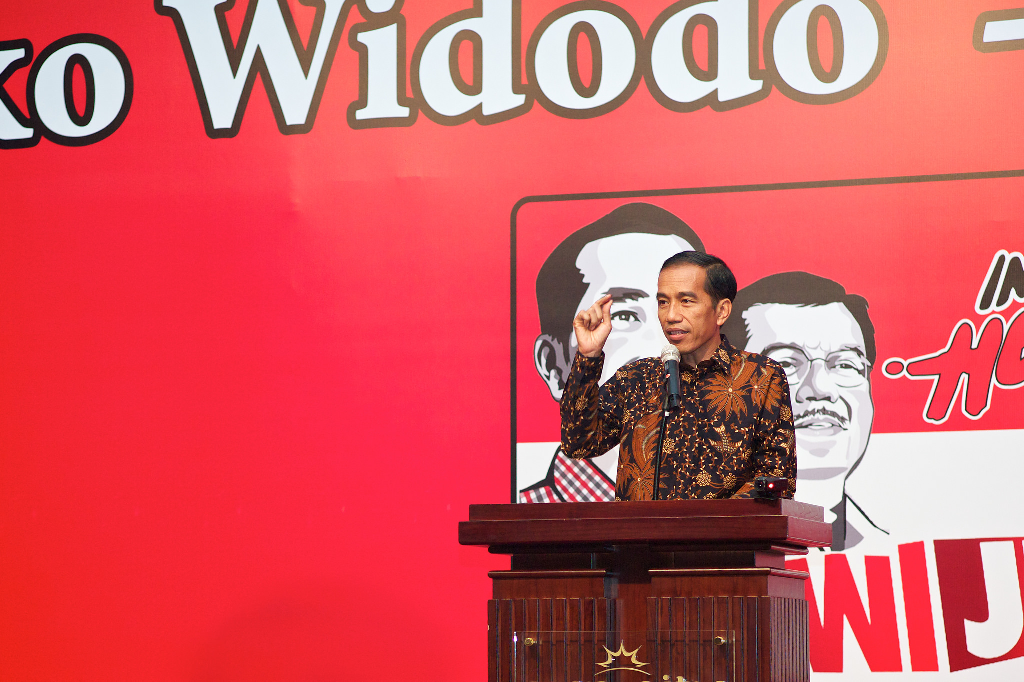 President Joko Widodo speaks at a podium, with a red banner as a backdrop.