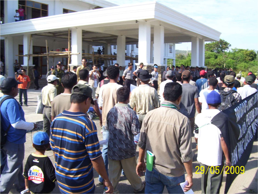 A group of people stand in front of a heavily guarded building, carrying a protest banner.