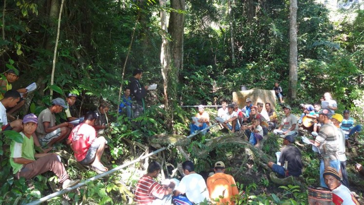 In a forest clearing, a group of villagers surround a priest, listening to his sermon.